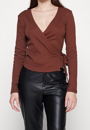Mujer vistiendo una blusa envolvente de manga larga marrón atada a un lado, combinada con pantalones de cuero negros de talle alto.
