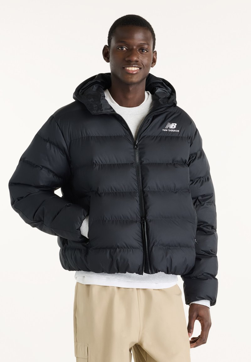 Homme souriant portant une doudoune noire New Balance avec capuche, une chemise blanche et un pantalon beige, debout devant un fond clair uni.