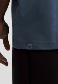 Kék pamut póló, lekerekített aljjal, az alsó bal sarokban egy kis logócímkével. Fekete nadrággal viselve, lazább szabásban.