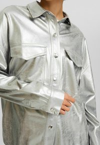 Silverfärgad metallisk skjortjacka med två bröstfickor, spetsig krage och tryckknappsstängning. Slät textur och avslappnad passform.