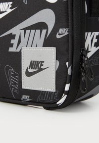 Sort Nike taske med hvide logo-tryk, fremstillet i et struktureret stof, med lynlåslukning og et sølv logo-patch på en rektangulær baggrund.
