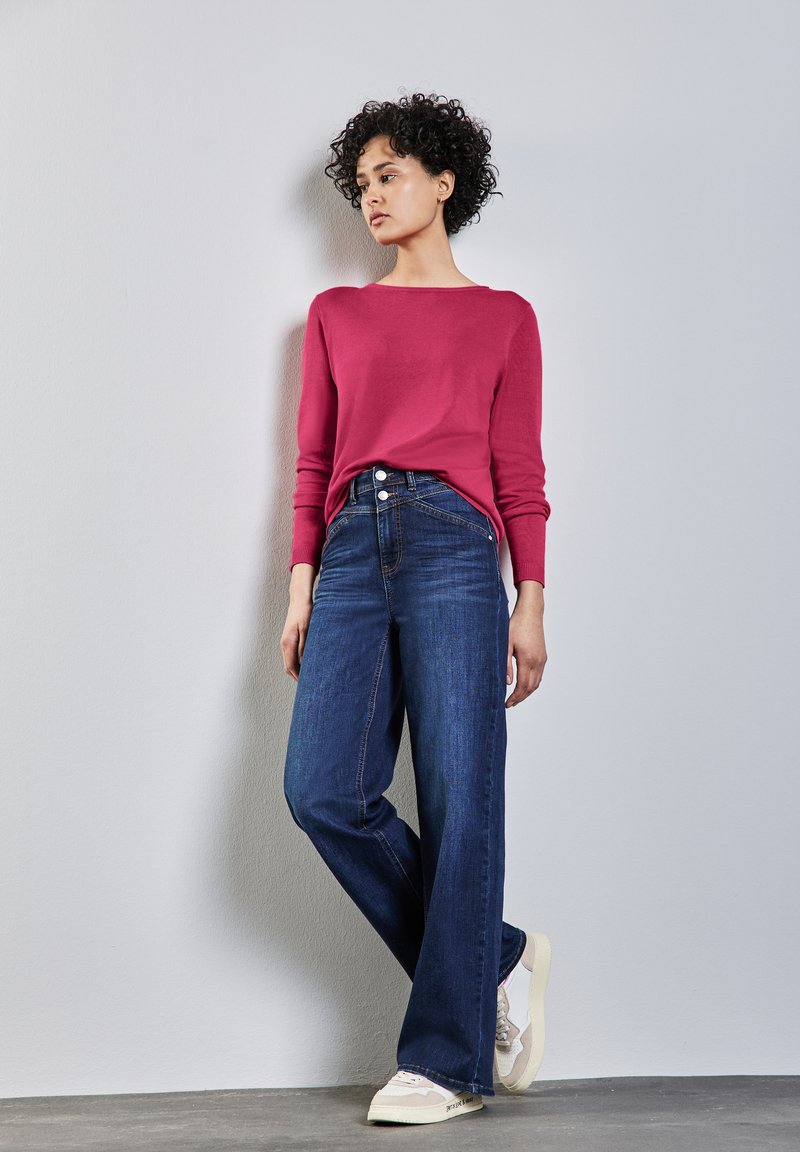 Enganfter rosa Pullover kombiniert mit weit geschnittenen dunkelblauen Jeans, die eine hohe Taille und einen Fünfknopfverschluss haben, ergänzt durch hellfarbene Sneakers.
