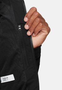 Sort Mammut DRI Tour jakke med mesh tekstur, tæt på et lynlås-lomme og label med syningsdetaljer.