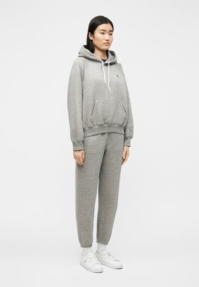 Γυναίκα που στέκεται φορώντας γκρι ανοιχτό φούτερ με κουκούλα, ταιριαστά παντελόνια τζόγκινγκ, λευκές κάλτσες και λευκά αθλητικά παπούτσια, μπροστά σε απλό φωτεινό φόντο.