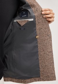 De voering van het bruin-zwart geruite blazer toont een gladde grijze stof. Bevat een zak en een "SLIM FIT" label, met een knoop zichtbaar.