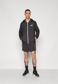 Rompevientos negro con capucha, cremallera blanca y logo de Puma. Combinado con pantalones cortos negros y zapatillas deportivas negras. Las piernas están tatuadas.