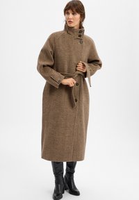 Frau passt ihren braunen Hahnentrittmantel mit hohem Kragen an, getragen zu schwarzen, kniebedeckenden Stiefeln mit Absatz, vor einem weißen Hintergrund.