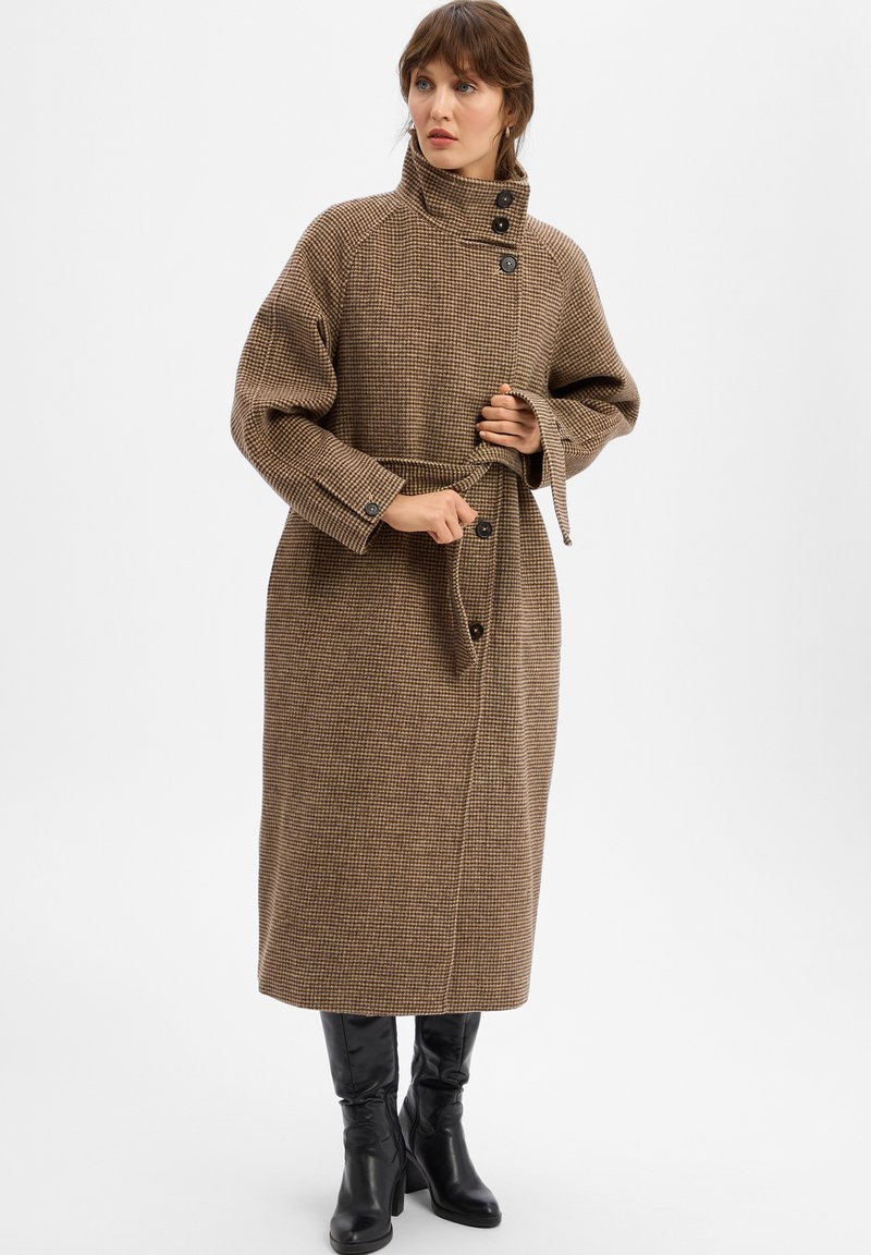 Frau passt ihren braunen Hahnentrittmantel mit hohem Kragen an, getragen zu schwarzen, kniebedeckenden Stiefeln mit Absatz, vor einem weißen Hintergrund.