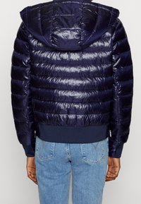 Granatowa kurtka puchowa o błyszczącym, pikowanym wykończeniu, z kapturem i ściągaczami na rękawach. Posiada kieszenie na zamek i dopasowany krój.