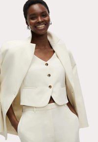 Wit op maat gemaakt driekstuk pak met een getailleerde blazer, een v-hals top met drie knopen, en bijpassende broek, allemaal in een gladde stof.