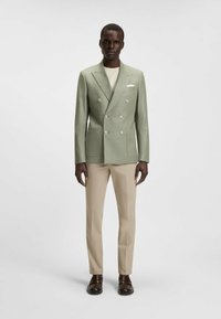 Lichtgroene dubbel onderhoud blazer met grote knopen, twee zakken; gecombineerd met beige broeken en donkerbruine schoenen. Vlekkeloze stofstructuur.