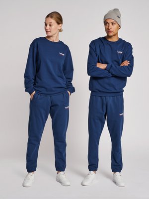 Mężczyzna i kobieta stoją, ubrani w pasujące granatowe bluzy i spodnie oraz białe trampki; mężczyzna nosi szarą czapkę, kobieta ma ręce w kieszeniach.