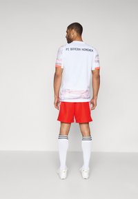 Fehér FC Bayern München mez pink és szürke részletekkel, piros nadrággal, fehér zoknikkal, fekete csíkokkal, valamint cipővel párosítva.