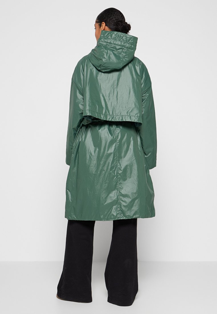 Parka verde, brillante, impermeable, con capucha, de diseño cropped y cinturilla ajustable. Combinado con pantalones anchos negros.