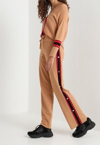 Beżowy sweter z dzianiny z dekoltem w serek i czerwonym paskiem akcentującym, zestawiony z dopasowanymi beżowymi spodniami z czerwonymi paskami po bokach i zatrzaskami. Czarne trampki.