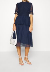 Navyblaues Kleid mit kurzen Spitzärmeln, plissiertem Rock und gestuftem Design. Kombiniert mit einer weißen Handtasche im Krokodildruck und Riemchenheels.