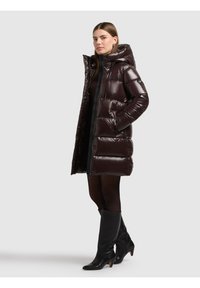 Sieviete, kas valkā spīdīgu tumši brūnu siltinātu kapuci ar melnu kleitu, zeķbiksēm un melnas ceļa augstuma zābakus ar papēžiem, stāv sānis.