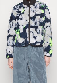 Kurtka polarowa w kolorach niebieskim, granatowym i szarym z neonowozielonymi akcentami. Posiada zamek błyskawiczny z przodu i małą kieszeń na piersi.