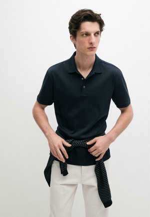 Jeune homme aux cheveux courts et foncés portant un polo noir à manches courtes, nouant un pull rayé autour de sa taille, debout devant un fond uni.