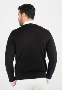 Pull en tricot noir à manches longues, col rond et poignets côtelés. Texture lisse avec un design ajusté, porté sur une chemise bleu clair.