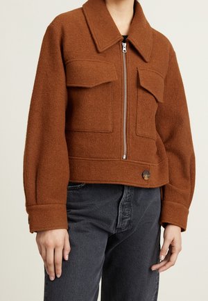 Henkilö, joka pukeutunut ruosteenväriseen villatakkiin, jossa on iso kaulus, vetoketju edessä, läppätaskut ja nappikoriste tummansinisten farkkujen päällä.