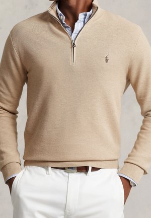 Mężczyzna w beżowym, teksturowanym swetrze z zamkiem do połowy długości, na niebieskiej koszuli w paski, białych spodniach i brązowym pasku, z rękami w kieszeniach.