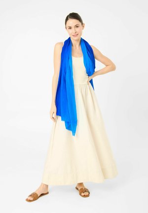 Vrouw draagt een crèmekleurige mouwloze jurk, een felblauwe sjaal om haar nek gedrapeerd en bruine open-toe sandalen, staand tegen een witte achtergrond.