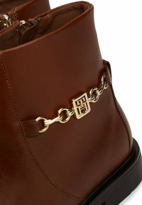 Bottine en cuir marron avec chaîne dorée et détail logo carré, fermeture éclair visible, et semelle épaisse noire.