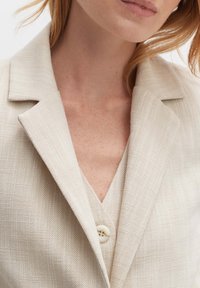 Blazer beige con finitura testurizzata, caratterizzato da un colletto a risvolto e un dettaglio con bottone. Il tessuto presenta una sottile trama diagonale.