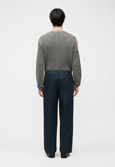 Άνδρας που στέκεται με την πλάτη προς τα εμπρός, φοράει υφασμένο γκρι πουλόβερ με μακριά μανίκια, σκούρο μπλε παντελόνι με πίσω τσέπες και μαύρα παπούτσια σε απλό φόντο.