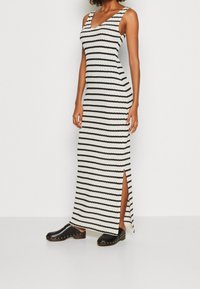 Femme portant une robe maxi blanche sans manches avec des rayures noires horizontales et une fente sur le côté, associée à des sabots noirs à enfiler ornés de clous dorés.