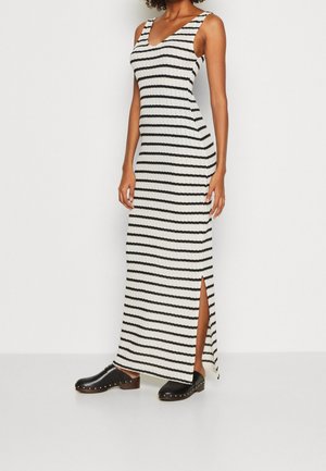 Vrouw in een mouwloze witte maxi-jurk met horizontale zwarte strepen en een zijsplit, gecombineerd met zwarte instapschoenen met gouden studs.