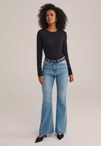 Kvinna med lockigt hår iklädd en svart långärmad tröja, ljusblå högmidjade utställda jeans, svart bälte och svarta spetsiga skor som står på en beige bakgrund.