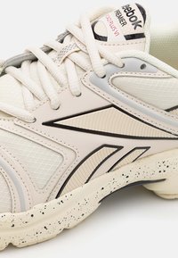Närbild av beige Reebok Premier Road Plus VI-sneakers med svarta detaljer, texturerade paneler och prickig sula.