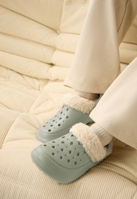 Ljusgröna träskor med en fluffig krämfärgad krage, med ventilationshål på ytan, bärs med beige byxor och ljusa strumpor.