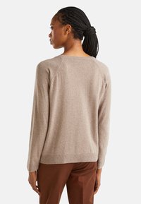 Kobieta z warkoczami, ubrana w beżowy sweter i brązowe spodnie, stojąca tyłem na tle jednolitego koloru.