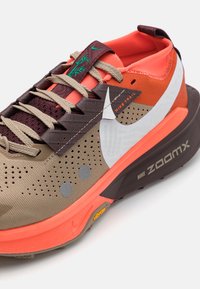 Nike ZoomX trail čevlji z zgornjim delom iz bež mreže, oranžnimi akcenti, sivimi pikami in debelim črnim podplatom z logotipom Vibram.