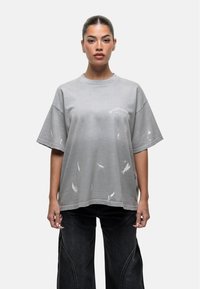 Szara, oversize koszulka z wystrzępionymi detalami i okrągłym dekoltem. Posiada drukowany tekst w kolorze białym. Materiał ma miękki, znoszony wygląd.