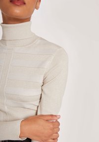 Lichtbeige coltrui met horizontale strepen, voorzien van een geribde hals en zachte, gestructureerde stof. Subtiele glans versterkt het ontwerp.