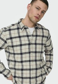 Geruit button-up overhemd in beige, zwart en bruin. Het heeft een kraag, lange mouwen en een relaxte pasvorm met een geruit patroon.