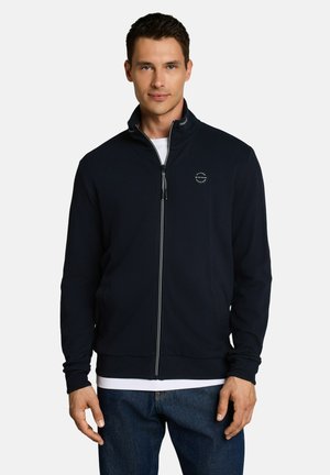 Mann trägt eine schwarze Zip-up-Jacke über einem weißen T-Shirt und blauen Jeans, steht vor einem einfarbigen hellen Hintergrund.