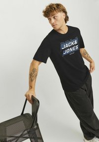 Sort bomulds-t-shirt med et stort blåt og hvidt "JACK & JONES" grafik. Modellen holder en sort mesh-stol og bærer mørke bukser.