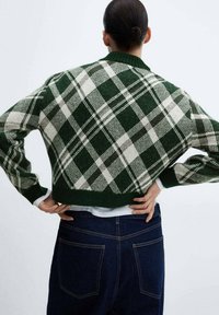 Pull court à carreaux verts et blancs avec un col arrondi et des poignets côtelés, porté avec un jean bleu foncé. Motif en tricot texturé bien visible.