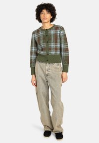 Zöld kockás kardigán kék és bordó mintákkal, kerek nyakkal és gombos elülső részel. Laid-back beiget farmernadrággal és fekete cipőkkel párosítva.