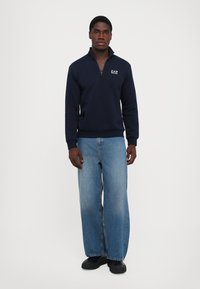 Marinegenser med kvart glidelås, med "EAZ"-logoen. Kombinert med lyseblå vide jeans og svarte joggesko. Flate, glatte teksturer.