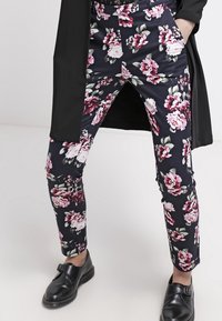 Květinové kalhoty z černé látky, s bílým a růžovým květinovým vzorem. Úzký střih s předními kapsami a manžetami na kotník. Černé boty.