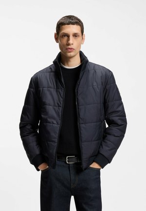 Młody mężczyzna w ciemnoniebieskiej pikowanej kurtce na czarnym swetrze i ciemnych jeansach, stojący z rękami w kieszeniach na jednolitym tle.