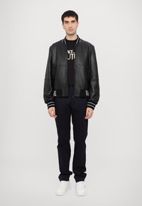 Jachetă bomber din piele neagră, cu manșete și tiv alb-striate, purtată peste un tricou grafic negru și pantaloni bleumarin, asociată cu adidași albi.