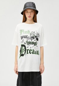 Camiseta blanca de gran tamaño con texto gráfico en verde y negro, diseños florales y un ajuste casual. Combinada con un sombrero de cubo negro.