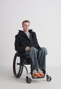 Schwarzer Mantel über einem grauen Pullover, blaue Jeans und orangefarbene Turnschuhe. Die Person sitzt in einem modernen Rollstuhl mit sleekem Design und großen Rädern.